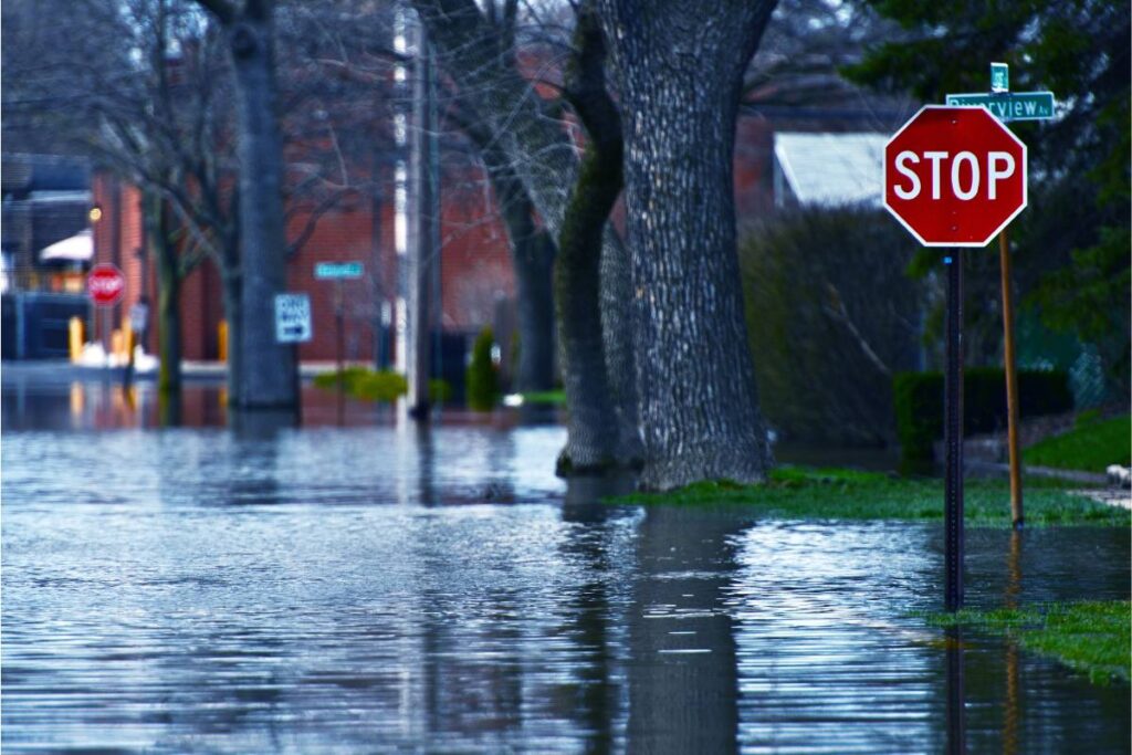A flooded street with a stop sign in focus, water submerging the roadway and lawns around residential buildings.